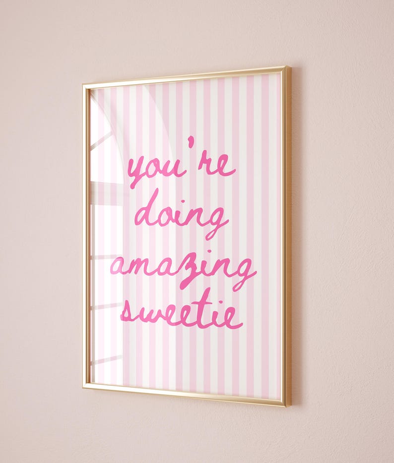 May include: A framed print with a gold-colored border. The artwork features a pink and white striped background with the handwritten phrase "you're doing amazing sweetie" in bright pink lettering.