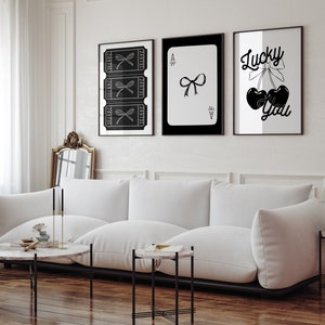 May include: Three black and white framed prints on a white wall. The left print features a series of tickets with a bow on each. The middle print is a black and white playing card, the Ace of Spades, with a bow on it. The right print features a black and white illustration of two cherries with a bow on top and the text "Lucky You".