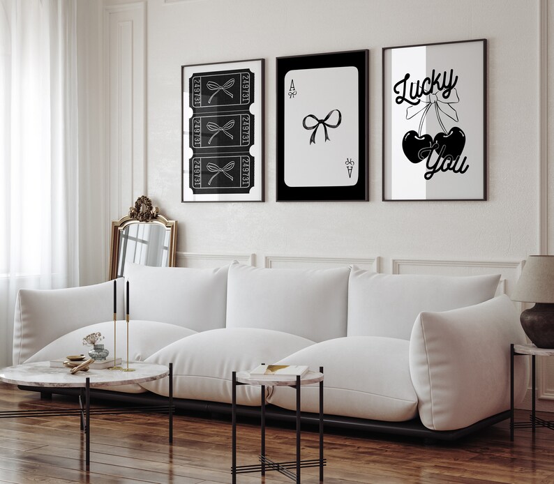 May include: Three black and white framed prints on a white wall. The left print features a series of tickets with a bow on each. The middle print is a black and white playing card, the Ace of Spades, with a bow on it. The right print features a black and white illustration of two cherries with a bow on top and the text "Lucky You".