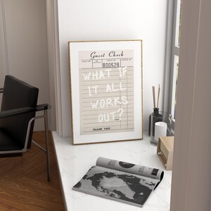 Puede incluir: Una impresi&oacute;n enmarcada de un "Guest Check" vintage con el texto "WHAT IF IT ALL WORKS OUT?" en blanco. Una revista enrollada con un mapa en blanco y negro est&aacute; frente al marco. Tambi&eacute;n se ven una vela, un difusor y una radio.