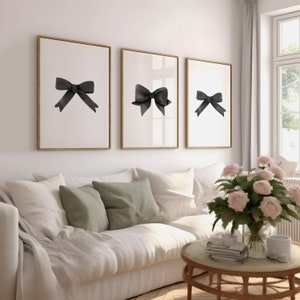 K&ouml;nnte beinhalten: Drei gerahmte Schwarzwei&szlig;-Aquarell-Illustrationen von Schleifen an einer wei&szlig;en Wand &uuml;ber einem wei&szlig;en Sofa mit gr&uuml;nen Kissen.