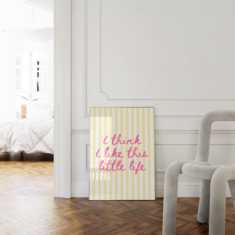 Puede incluir: Un p&oacute;ster enmarcado con un fondo a rayas amarillas y blancas y el texto "I think I like this little life" en escritura rosa. El p&oacute;ster est&aacute; apoyado contra una pared blanca junto a una silla gris.