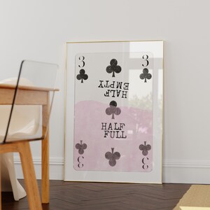 Puede incluir: Impresión artística enmarcada con un diseño de carta de juego. La carta es un tres de tréboles, con las palabras "HALF EMPTY" y "HALF FULL" impresas. El fondo tiene un degradado rosa. Marco dorado.