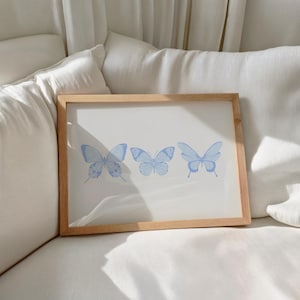 Puede incluir: Una impresión enmarcada con tres ilustraciones de mariposas azules sobre un fondo blanco. Las mariposas están dispuestas horizontalmente, cada una con un patrón de alas único. El marco es de madera clara.