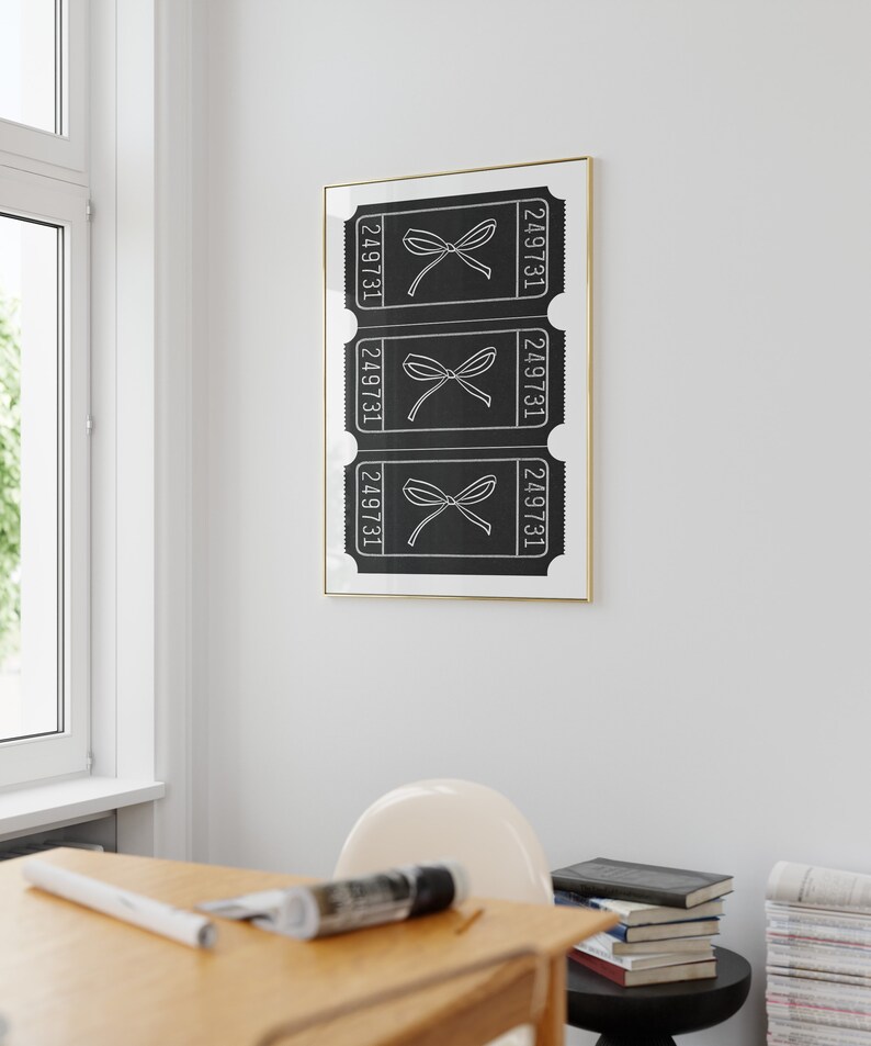 May include: Black and white print of three ticket stubs with the number 249731 on each. The tickets have a bow on them. The print is framed in gold and hanging on a white wall.