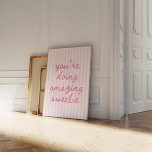 May include: A framed pink and white striped print with the words "you're doing amazing sweetie" in pink script. The print leans against a white wall next to a gold-framed canvas. The room has a wooden floor.