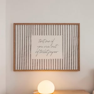 Puede incluir: Un cuadro rectangular con fondo de tela clara y rayas grises verticales, enmarcado en madera. En el centro, un papel color crema con bordes festoneados muestra el texto en cursiva negra: text me if you run out of toilet paper.
