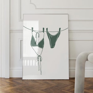 Peut inclure: Une affiche encadrée présentant une illustration minimaliste d'un bikini vert, composé d'un haut et d'un bas, suspendu à une corde à linge. L'œuvre est posée sur un fond blanc, avec une esthétique épurée. L'affiche est exposée dans une pièce avec un parquet.