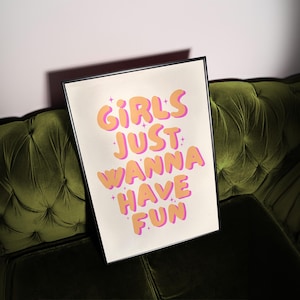 Puede incluir: Una impresión enmarcada con el texto "Girls Just Wanna Have Fun" en un esquema de color rosa y naranja. El texto está estilizado con una fuente redondeada y burbujeante.