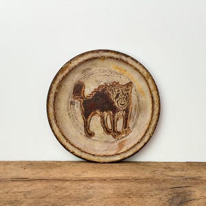 May include: A circular ceramic plate in brown and cream tones. The plate has a carved design of a stylised cat in the centre. The cat is brown, and the plate has a textured, rustic appearance.