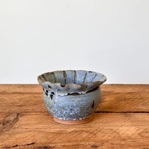 May include: A small, handmade ceramic bowl with a blue-gray glaze. The bowl has a ruffled rim and decorative cutouts. It sits on a small, unglazed terracotta base. The bowl is displayed on a rustic wooden surface.