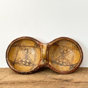 May include: A double-bowl, hand-painted, antique-style wooden bowl. The bowl has a light brown interior with a geometric black design. The exterior is a darker brown with a reddish hue. The bowl sits on a wooden surface.