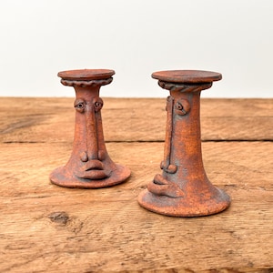 May include: Two terracotta-coloured ceramic candle holders with a textured, matte finish. Each piece features a sculpted face with prominent eyes, a long nose, and full lips on the central column, topped with a circular, rope-patterned rim.