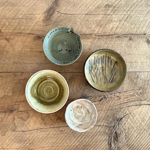 Puede incluir: Cuatro cuencos de cerámica hechos a mano en tonos verdes, beige y blanco están dispuestos sobre una superficie de madera. Los cuencos varían en tamaño y diseño, uno con asa y otro con un motivo de espadañas.