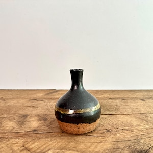 Puede incluir: Un pequeño jarrón de cerámica negra con cuello estrecho y cuerpo redondeado. Una banda dorada horizontal rodea la mitad del jarrón, sobre una base texturizada de color marrón claro. El jarrón está sobre una superficie de madera.