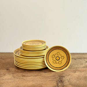May include: A collection of yellow plates featuring a brown and gold floral pattern. The plates are stacked, with one plate separate. The plates are displayed on a wooden surface. The plates are approximately 20 cm in diameter.
