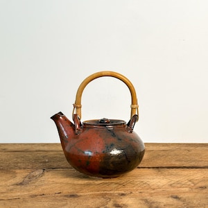 May include: A ceramic teapot with a bamboo handle. The teapot is a mix of red and black with a glossy finish. The spout and lid are visible. The teapot sits on a wooden surface.