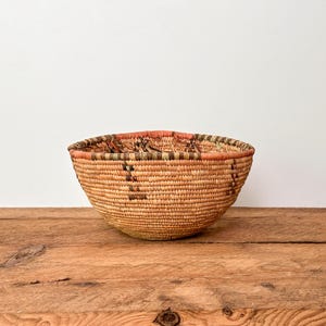 May include: A woven basket with a rounded form and a broad opening. It's crafted from natural fibres in tan and brown tones, with darker decorative patterns. The rim has an orange and green band. The basket is likely for storage.
