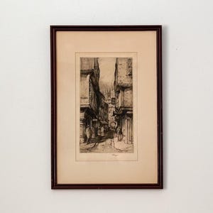 Puede incluir: Grabado en blanco y negro enmarcado de una escena callejera europea. La obra representa una calle estrecha con edificios a ambos lados y figuras a lo lejos. El marco es marrón oscuro.