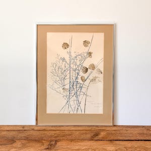May include: Framed botanical print with beige, blue, and gold stems and seed pods. The artwork is in a silver frame against a neutral backdrop, creating a minimalist aesthetic. The print is displayed on a wooden surface. The frame measures approximately 30 x 20 cm.