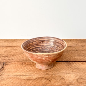 May include: A rustic, antique-style bowl with a wide rim and a footed base. The bowl features a reddish-brown exterior and a dark brown interior with a repeating pattern. The bowl is set on a wooden surface.