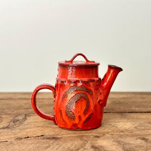 Puede incluir: Una tetera de cerámica roja con tapa y asa. La tetera tiene una superficie texturizada con detalles marrones. El pico y el asa son curvos. La tetera está sobre una superficie de madera.