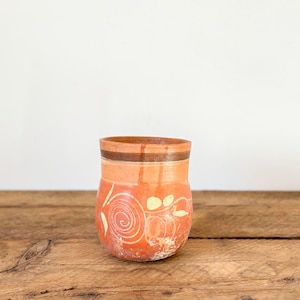 May include: A terracotta-coloured ceramic vase with a cylindrical shape. The vase has a dark brown band near the rim and is decorated with white spiral and leaf-like designs. It sits on a rustic wooden surface.