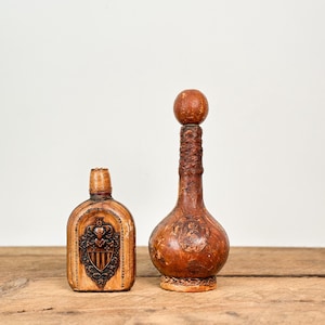May include: Two antique brown leather bottles. One is a small flask with a shield design. The other is a tall bottle with a bulbous base and a decorative top. Both bottles are on a wooden surface.