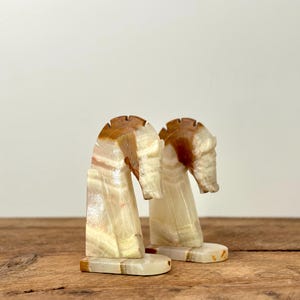May include: A pair of horse head bookends, carved from cream-coloured stone with brown and tan markings. The decorative items are displayed on a wooden surface. Each bookend is approximately 10 cm tall.