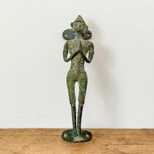 May include: An antique bronze figure of a person with hands clasped in prayer. The figure has a detailed design and is mounted on a decorative backplate. The figure is standing on a base with two holes.