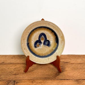 May include: A round ceramic plate featuring a speckled beige and tan glaze with a central dark blue abstract motif. The plate is displayed on a brown wooden stand, showcasing a smooth rim and a deep, circular basin with a glossy finish.