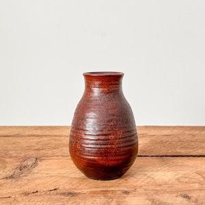 May include: A handcrafted, reddish-brown ceramic vase with a textured surface. The vase has a rounded body and a slightly flared rim. Horizontal ridges encircle the body. The vase sits on a wooden surface.