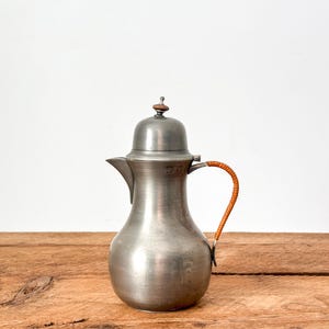 Puede incluir: Una jarra de peltre vintage con tapa y asa trenzada. La jarra tiene un cuerpo bulboso y un pico. El asa es de material trenzado marrón claro. La jarra está sobre una superficie de madera.