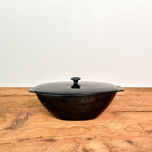 May include: A glossy black ceramic casserole dish with a matching lid and integrated side handles. The bowl has a rounded, deep basin shape and a smooth, reflective surface. The lid features a small, centered knob handle for easy removal.