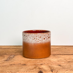 May include: A cylindrical ceramic planter featuring a smooth, solid terracotta-coloured base that transitions into a textured, bubbly white glaze around the upper rim. The interior of the vessel is finished in a deep, glossy reddish-brown glaze.