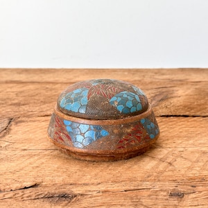 May include: A small, round antique box with a lid. The box is made of a dark brown material and is decorated with blue and red floral patterns. The box sits on a wooden surface.