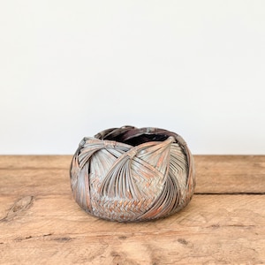 May include: A circular woven basket with an intricate, overlapping design. The basket is made from natural-coloured reeds with reddish-brown accents. It rests on a wooden surface against a white backdrop.