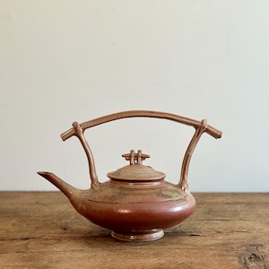 May include: A handcrafted, reddish-brown ceramic teapot with a curved handle and a spout. The lid features a small, decorative handle. The teapot sits on a wooden surface, showcasing its rustic charm and unique design. The teapot's dimensions are approximately 25 cm wide and 20 cm tall.