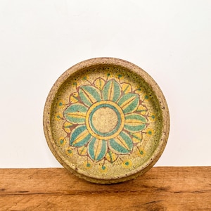 May include: A round ceramic dish with a speckled beige and brown textured rim. The centre features a hand-painted floral motif with teal petals outlined in dark brown, surrounded by yellow accents and small dots on a light yellow background.
