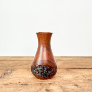 May include: A small, handcrafted wooden vase with a bulbous base and a narrow neck. The vase is made of a rich, reddish-brown wood, with a textured, dark brown section near the base. The vase sits on a wooden surface.