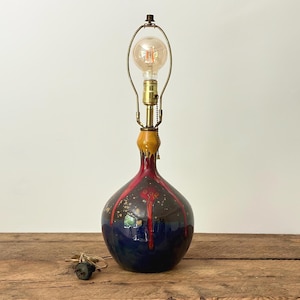 Peut inclure: Une lampe de table en céramique avec une base bleu foncé, ornée d'accents rouges et dorés. La lampe présente un col en laiton et une ampoule de style vintage. La lampe est posée sur une surface en bois.