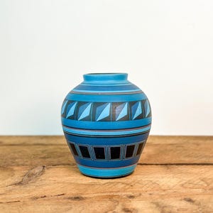 May include: A blue ceramic vase with a geometric design. The vase features horizontal bands of varying shades of blue, with black and white geometric shapes. The vase is sitting on a wooden surface.