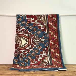 May include: A rectangular, patterned rug with a blue background and a complex design in red, white, and orange. The rug features geometric shapes and floral motifs, with a border of repeating patterns. The rug is draped over a white rod.