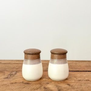 Puede incluir: Dos saleros y pimenteros de cerámica con tapa marrón, banda marrón claro y base blanca. Los saleros y pimenteros están sobre una superficie de madera con fondo blanco.
