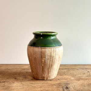 May include: A decorative vase with a green glazed top and a natural, woven base. The vase has a rounded shape and sits on a wooden surface. The green top contrasts with the light brown woven texture of the base.