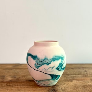 May include: A round, pink ceramic vase with a teal, marbled design. The vase sits on a wooden surface against a white background. The teal pattern swirls around the vase, creating a unique decorative piece.