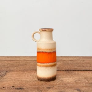 Puede incluir: Un jarrón de cerámica vintage con asa, con un cuerpo color crema con bandas horizontales naranjas, marrones y beige. El jarrón mide aproximadamente 25 cm de alto y está sobre una superficie de madera.