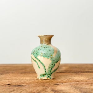 May include: A small, handcrafted ceramic vase with a beige neck and base, and a cream-coloured body. The vase is adorned with a textured, speckled green glaze that drips down the sides. The vase is approximately 15 cm tall.