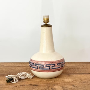 Puede incluir: Lámpara de mesa de cerámica color crema con base bulbosa y cuello delgado. La base presenta una banda decorativa de patrones geométricos azules y rojos. La lámpara tiene una parte superior de color latón y un cable blanco.
