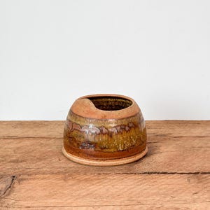 May include: A white ceramic mug with a black handle and the text "But First, Coffee" in a bold, black font. The mug is set against a blurred background, suggesting a cosy setting.